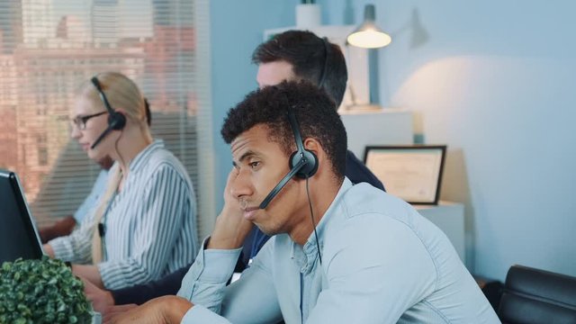 Bored multiethnic customer support operator waiting for telephone call from client. His colleagues are busy working.