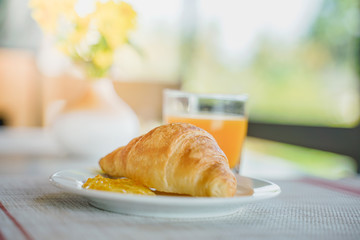croissant on white plate with orange marmalade and juice on table in morning, soft focus