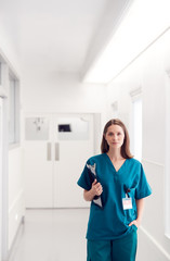 Portrait Of Female Doctor Wearing Scrubs Standing In Hospital Corridor Holding Clipboard