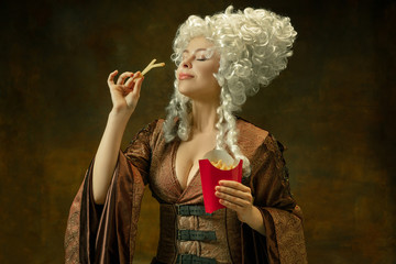 Astonished eating fried potato. Portrait of medieval young woman in vintage clothing on dark background. Female model as a duchess, royal person. Concept of comparison of eras, modern, fashion, beauty