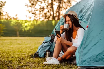 Schilderijen op glas Portret van een jonge vrouw op de camping, zittend aan een tent koffie drinkend en kijkend naar een smartphone. © bnenin