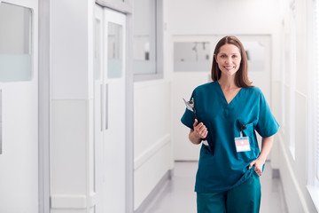 Portrait Of Smiling Female Doctor Wearing Scrubs Standing In Hospital Corridor Holding Clipboard