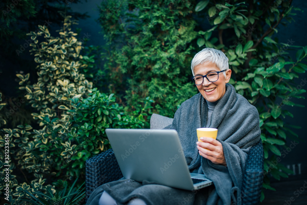 Poster Portrait of a cheerful modern elderly woman using laptop in the garden.