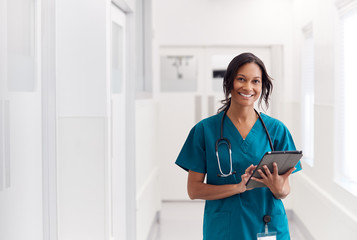 Portrait Of Smiling Female Doctor Wearing Scrubs In Hospital Corridor Holding Digital Tablet - obrazy, fototapety, plakaty