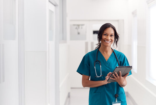 Portrait Of Smiling Female Doctor Wearing Scrubs In Hospital Corridor Holding Digital Tablet