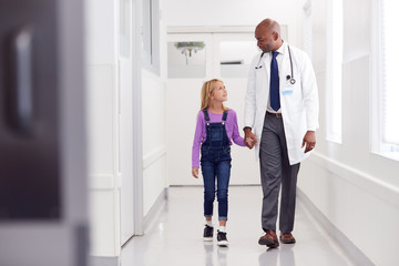Male Paediatric Doctor Walking Along Hospital Corridor Holding Hands With Young Girl Patient
