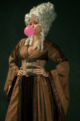 Pink bubble gum. Portrait of medieval young woman in brown vintage clothing on dark background....