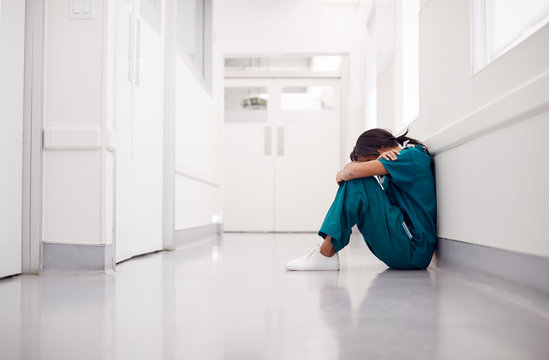 Stressed And Overworked Female Doctor Wearing Scrubs Sitting On Floor In Hospital Corridor
