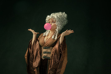 Pink bubble gum. Portrait of medieval young woman in brown vintage clothing on dark background. Female model as a duchess, royal person. Concept of comparison of eras, modern, fashion, beauty.
