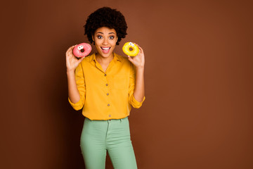Photo of funny pretty dark skin curly lady hold two colorful donuts with caramel eyes human faces good mood wear yellow shirt green pants isolated brown color background