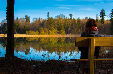 Mujer sentada en un banco mirando al reflejo del bosque en el lago