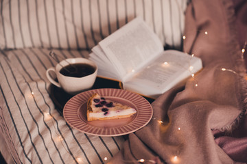 Open book, cup of black tea and tasty fruit cake on wooden tray in bed closeup over glowing lights at background. Good morning. Breakfast time.