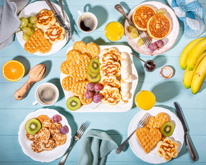 Healthy breakfast with fresh hot waffles hearts, pancakes flowers with berry jam and fruits on turquoise background, top view, flat lay. Food concept.