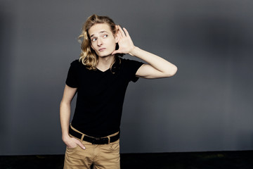 Attractive guy with long blonde hair, substituting a hand to his ear, eavesdrops with an interested...
