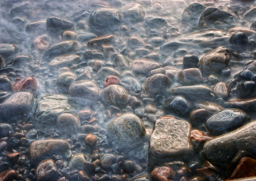 Bunte, runde, abgeschliffene Steine liegen am Strand in der Sonne und werden vom Wasser umsp&uuml;lt