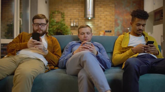 Gadget addicted young men sitting on couch with smartphones, boring home party