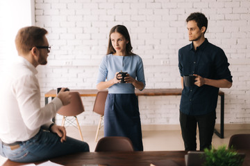 Business team sharing ideas during coffee break. Two handsome young businessmen and lady in casual clothing are holding cups of coffee, talking and smiling, standing indoors the office building