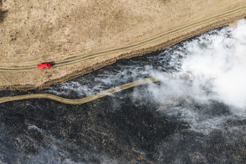 Field fire and red fire truck near firebreak, aerial top down view. Dry grass burning in countryside with gray smoke, black burnt land remains