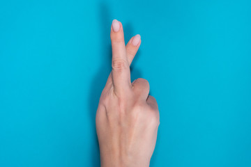 Closeup top view photography of white female hand making gesture with two crossed fingers as symbol of good luck. Hand isolated on bright blue background.