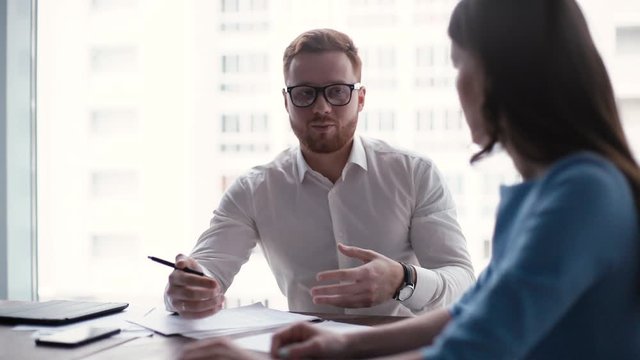 Two colleagues man and woman have business communication in office against the window. Stylish guy with glasses talks about his opinion on the new project. Tracking shot in slow motion.