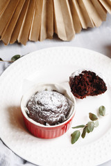 Chocolate muffins in red cups. Small glazed ceramic ramekin with brown cakes on a gray and white background.