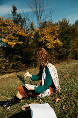 Portrait of young female artist sitting and working on painting outdoors, in the autumn landscape. She is in front of the canvas and drawing.She holds oil paints, artist brushes, canvas and palette. 