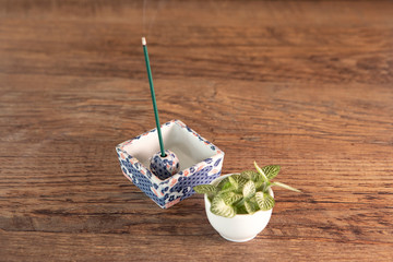 Arrangement with incense stick burning in beautiful ceramic incense holder, wooden table background with copy space, some fresh green plant aside. 