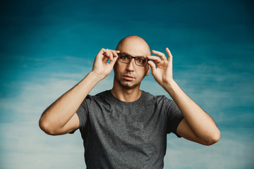 Naklejka premium An interesting bald man adjusts his glasses with his hands and looks in surprise at the camera on a blue background.