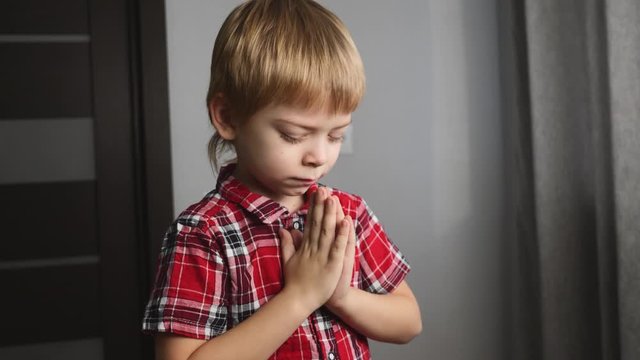 A blond boy prays alone in his room. The child holds his hands together.