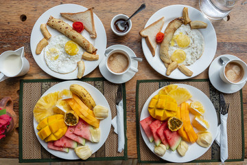 Tropical breakfast of fruit, coffee and scrambled eggs and banana pancake for two on the beach near sea. Top view, table setting.