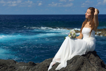 Fototapeta na wymiar Elegant bride in light white wedding dress sitting on ocean rock shore, flowers in hand. Wedding ceremony on ocean coast
