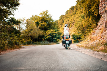 Wedding, newlyweds. A man and a woman in wedding dresses and motorcycle helmets, riding a motorcycle on the road. The woman stands. Copy space. Horizontal. Outdoor