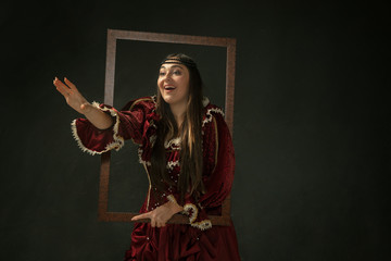 Self portrait. Portrait of medieval young woman in red vintage clothing standing on dark background. Female model as a duchess, royal person. Concept of comparison of eras, modern, fashion, beauty.