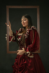 Proud. Portrait of medieval young woman in red vintage clothing standing on dark background. Female model as a duchess, royal person. Concept of comparison of eras, modern, fashion, beauty.