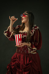 Watching cinema. Portrait of medieval young woman in red vintage clothing standing on dark...