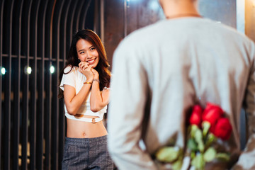 Asian man hiding roses flower to surprise his girlfriend on dating in Valentine's, Anniversary or birthday, focus on stanging happy woman smile on face, attractive couple having romantic on rooftop