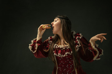 Passioned. Portrait of medieval young woman in red vintage clothing eating burger on dark background. Female model as a duchess, royal person. Concept of comparison of eras, modern, fashion, beauty.