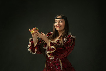 First love. Portrait of medieval young woman in red vintage clothing holding burger on dark...
