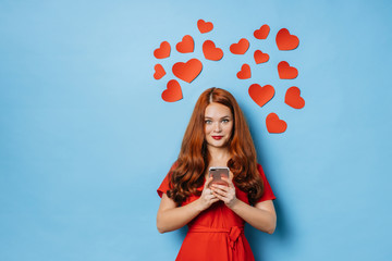 redhaired woman in love, female in red dress stand isolated over blue background, hold smartphone...