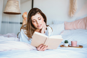 Girl reading a book, drinking coffee or tea, resting in bed.
