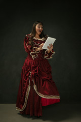 Astonished. Portrait of medieval young woman in red vintage clothing using tablet on dark background. Female model as a duchess, royal person. Concept of comparison of eras, modern, fashion, beauty.