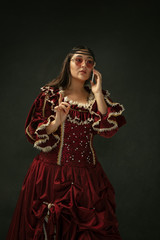 Talks on phone wearing modern eyewear. Medieval young woman in red vintage clothing on dark...