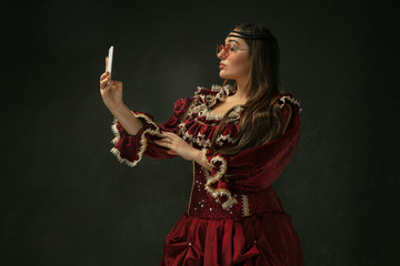 Takes selfie wearing modern eyewear. Medieval young woman in red vintage clothing on dark background. Female model as a duchess, royal person. Concept of comparison of eras, modern, fashion, beauty.
