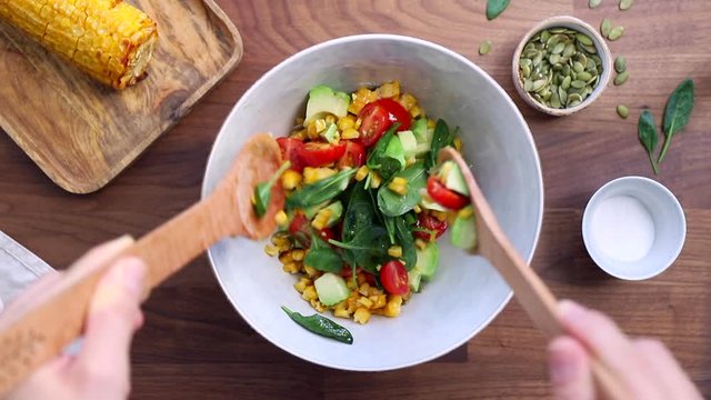 Hands mix summer vegetable salad with spinach, cherry tomatoes, avocado, grilled corn and pumpkin seeds. Healthy eating concept, top view.