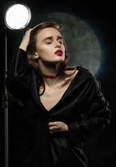 Beautiful slim braless girl, wearing a unbuttoned black blazer, sensually frowns and touches her hair with her hand on a dark background, posing next to a light lamp. Advertising, trendy design.