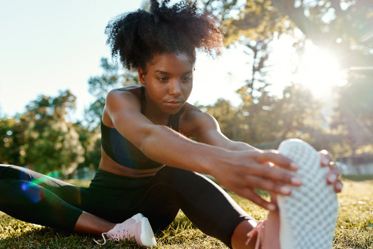 Portrait of an african american young woman sitting in the park stretching her legs in bright sunlight - young black woman warming up her legs before doing exercise
