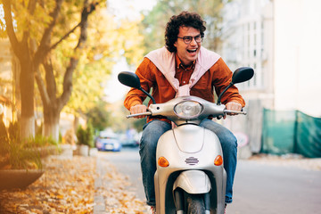 Outdoor image of smart young man driving on his scooter, dressed in casual outfit and transparent eyeglasses, with curly hair, has joyful expression.