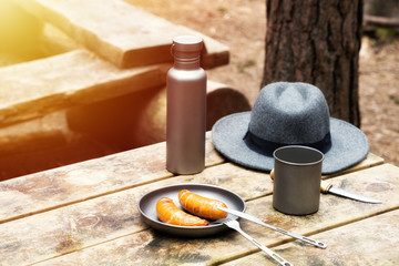 Knife, hat, water bottle and titanium mug on the wooden table. Grilled sausages on the plate. Top view. Outdoors. Bushcraft concept.