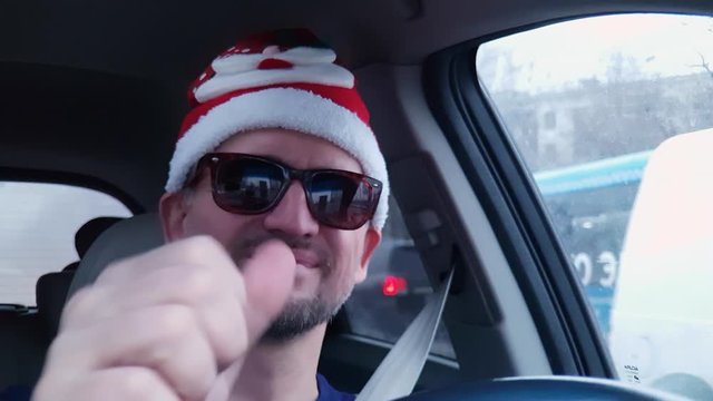 White bearded man in red Santa Claus hat and sunglasses with smile drives passenger car and waves at camera