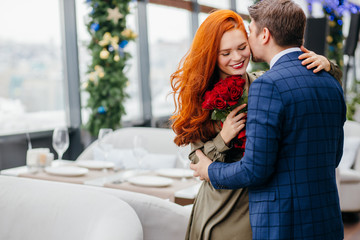 kind and gallant caucasian man in tuxedo invited on a dtae and gave red flowers, roses to his lady in dress. smiling pleasant redhaired woman thanks him. in restaurant